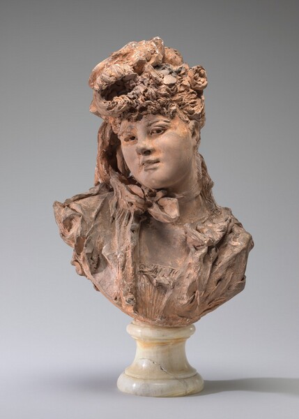 The head and shoulders of a young woman with an elaborate updo is shown in this free-standing terracotta bust. Her shoulders face us and her head tips to our left in this photograph. She looks at us from the corners of her eyes under low brows. She has a snub nose, her bow-shaped lips are parted, and she has smooth, full cheeks in her round face. Curls are piled atop her head and some rest low on her forehead. Her hair along with a flower to one side and a suggestion of a hat are indistinct because of the loose sculpting here. More hair falls down the right side of her face, to our left, and down the nape of her neck. A bow is tied like a choker and ruffles suggest a bodice and perhaps a coat. The bust ends in an upward curve and rests on a polished, white stone pedestal.
