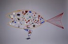 The outline of a fish is made with bent, colored wire and then hung with fragments of colored glass to loosely resemble scales in this free-floating sculpture. In this photograph, the open mouth faces our left. The body is made with a thick gauge, mustard-yellow wire. Finer, gold-colored wire creates a honeycomb-like pattern within the body. Pieces of colored glass hang within most of the cells. The irregularly shaped pieces are clear or in tones of brick red, indigo or sky blue, amber brown, pearl white, or emerald green. A few cells have little bits of metal, including a washer, a disk pierced with holes, and a spiraling piece of wire. The thicker yellow wire makes a circle near the mouth, creating an eye, which is hung with two pieces of glass in plum purple and pale lilac. To our right, the tail is as tall as the body and half its length. It is outlined with thick, red wire. Another piece of wire runs along the middle of the tail, and thinner red wire is strung from the central wire to the upper and lower edges, mimicking the rays of the fish’s fin. A fin along the bottom center of the fish’s body is made with thick, royal-blue wire that balances across the bottom rim of the fish. On the side closer to us, the blue wire is bent into a boomerang shape. It is suspended within a cell close to the bottom of the fish’s body, and is balanced on the far side with a piece of cobalt-blue glass. A hand-shaped pendant hangs from the point of the fin closest to us. The wall behind the hanging sculpture is fog gray.