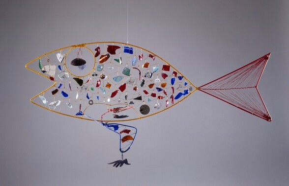 The outline of a fish is made with bent, colored wire and then hung with fragments of colored glass to loosely resemble scales in this free-floating sculpture. In this photograph, the open mouth faces our left. The body is made with a thick gauge, mustard-yellow wire. Finer, gold-colored wire creates a honeycomb-like pattern within the body. Pieces of colored glass hang within most of the cells. The irregularly shaped pieces are clear or in tones of brick red, indigo or sky blue, amber brown, pearl white, or emerald green. A few cells have little bits of metal, including a washer, a disk pierced with holes, and a spiraling piece of wire. The thicker yellow wire makes a circle near the mouth, creating an eye, which is hung with two pieces of glass in plum purple and pale lilac. To our right, the tail is as tall as the body and half its length. It is outlined with thick, red wire. Another piece of wire runs along the middle of the tail, and thinner red wire is strung from the central wire to the upper and lower edges, mimicking the rays of the fish’s fin. A fin along the bottom center of the fish’s body is made with thick, royal-blue wire that balances across the bottom rim of the fish. On the side closer to us, the blue wire is bent into a boomerang shape. It is suspended within a cell close to the bottom of the fish’s body, and is balanced on the far side with a piece of cobalt-blue glass. A hand-shaped pendant hangs from the point of the fin closest to us. The wall behind the hanging sculpture is fog gray.