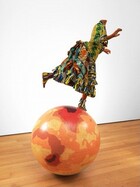 A headless mannequin of a person dressed in a colorful, patterned gown balances on a globe of the Earth in this free-standing sculpture. The person has medium-brown skin and, in this photograph, her body faces our right in profile. She balances on one foot, which is planted near the Arctic Circle, and her other foot is raised behind her, as if running. Her arms are lifted, palms facing outward. The bodice of her dress is mustard yellow with a swirling green and blue pattern running down the sleeve we can see. The cuffs and hem are trimmed with ruffles striped in navy blue, yellow, and white, some with thin, horizonal blue lines. The same fabric is gathered in horizontal pleats down the front of the dress. The rest of the skirt is a mix of maroon-red fabric decorated with geometric patterns in pumpkin orange, teal green, golden yellow, and ginger brown. The fabric is gathered up like a bustle at the girl’s lower back. Her ankle boots are made from the fabric patterned in blue, yellow, white, and maroon. Much of the globe is covered in areas of honey yellow and marigold orange. A few areas have darker red zones, including a rust-red band curving from Western to Southern Africa. Smaller, even darker zones are nested over the southern part of that continent. A zone of crimson red covers much of Iran and Afghanistan, and reaches into Russia and the surrounding areas, at the top of the globe. The sculpture sits on a hardwood floor and is shown against a white wall.