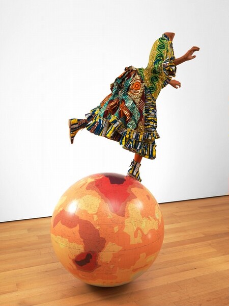 A headless mannequin of a person dressed in a colorfully patterned gown balances on a globe of the Earth in this free-standing sculpture. The person has medium-brown skin and, in this photograph, her body faces our right in profile. She balances on one foot, which is planted near the Arctic Circle, and her other foot is raised behind her, as if running. Her arms are lifted, palms facing outward. The bodice of her dress is mustard yellow with a swirling green and blue pattern running down the sleeve we can see. The cuffs and hem are trimmed with ruffles striped in navy blue, yellow, and white, some with thin, horizonal blue lines. The same fabric is gathered in horizontal pleats down the front of the dress. The rest of the skirt is a mix of maroon-red fabric decorated with geometric patterns in pumpkin orange, teal green, golden yellow, and ginger brown. The fabric is gathered up like a bustle at the girl’s lower back. Her ankle boots are made from the fabric patterned in blue, yellow, white, and maroon. Much of the globe is covered in areas of honey yellow and marigold orange. A few areas have darker red zones, including a rust-red band curving from Western Africa to Southern Africa. Smaller, even darker zones are nested over the southern part of that continent. A zone of crimson red covers much of Iran and Afghanistan, and reaches into Russia and the surrounding areas, at the top of the globe. The sculpture sits on a hardwood floor and is shown against a white wall.