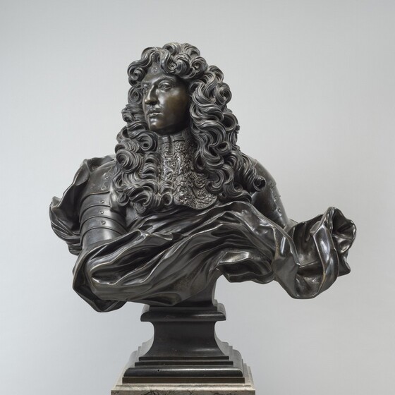 A smooth-skinned man is shown from the shoulders up amid a swirl of curls and fabric in this freestanding bronze sculpture. In this photograph of the piece, his shoulders square to us as he looks off to our left, his chin slightly lifted. He has bushy brows, a prominent nose, smooth cheeks, a pointed chin, and his full lips are closed. A thin mustache brushes his upper lip, and a curl coils in a perfect spiral on his somewhat flattened forehead. Curls cascade from head to well beyond his shoulders to flank a wide, patterned collar. We catch glimpses of armor on his shoulders within a whirl of billowing fabric. Below, a rectangular pedestal with a tall, inward-curving cove over stepped molding is on a marble plinth. The sculpture is shown against a fog-gray background in this photograph.