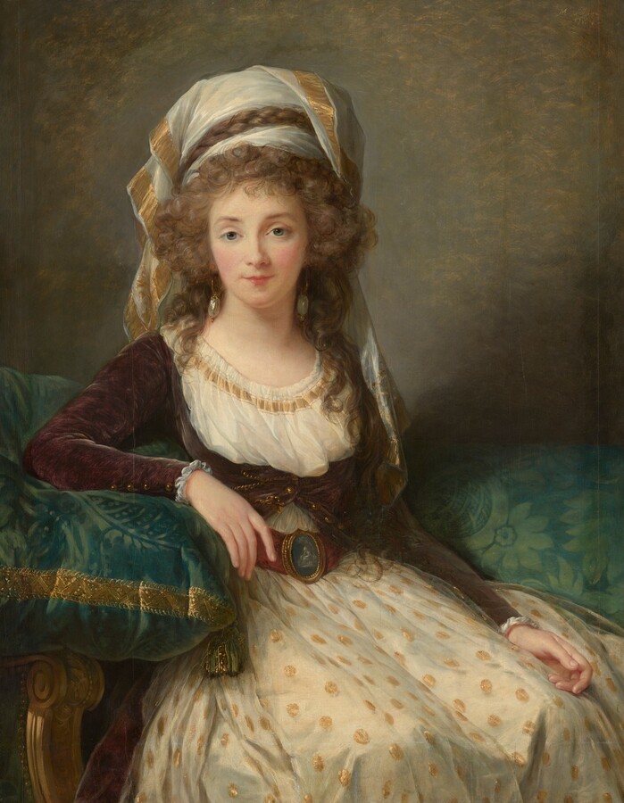 Shown from the knees up, a young woman with pale skin, wearing a short wine-red jacket over an ivory-white gown, gazes out at us from this vertical portrait painting. She sits in a chair surrounded by emerald-green cushions, which are covered with a light green floral pattern. The woman’s shoulders are squared toward us as her knees angle slightly to our right. The skirt of her gown is patterned with coin-sized gold dots against the white background, and the scooped neckline of her bodice is trimmed with gold ribbon. The top of at least four buttons on her velvet jacket are buttoned under her bust, and a cinnamon-brown belt with an iron-gray buckle, perhaps a cameo, cinches her waist. Her long, ash-brown hair is loosely pulled up with curls faming her face and falling to her shoulders. One braided plait is interwoven through a loose white turban, the ends of which drape over her shoulders. She has gray eyes under thin brows, a rose-red bow mouth, and her cheeks are tinged with pink. Gleaming, laurel-green drop earrings dangle from her ears. Her right arm, to our left, rests on an oversized green cushion, which balances on the carved, gold arm of her chair. Soft light from the upper left glints on the gold ribbons of the turban and cushion, and on her earrings and jacket buttons. The wall behind her is mottled with smoke gray and moss green. The artist has signed and dated the painting in the upper right, “Mde Le Brun 1789.”