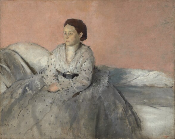 Shown from about the ankles up, a woman with ashen, pale skin, wearing a full, silvery-white dress with gray dots, sits on a low-backed white sofa against a pale peach background in this horizontal portrait painting. Just to our left of center, the woman sits with her body angled to our left and she looks in that direction with pale green eyes. Touches of green shade the inner corners of the eyes, and her complexion has a yellowish cast. Her pink lips are closed and her chestnut-brown hair is parted in the middle and pulled back. Her hands rest in her lap with wrists crossed. The dress darkens from eggshell white on the chest and sleeves to nickel gray on the skirt. Charcoal-gray dots create an irregular pattern that becomes looser as it moves down the skirt. Her dress is cinched at the waist with a black sash. The skirt spreads over her settee, which has a low back that angles up on our left. The peach wall behind her takes up the top half of the composition. The painting is created blended brushstrokes, giving it a soft, almost hazy look. The artist’s signature in red appears in the lower right corner: “Degas.”