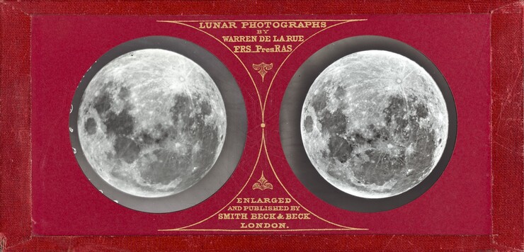 Two nearly identical black and white images of the lunar surface are encased in round openings in a rectangular, red holder. The edges of the holder are wrapped with tape or other material in a similar red color. The photographs are to either side of decorations and text in gold that create a vertical line up the center, between the photographs. Text along the top reads, “LUNAR PHOTOGRAPHS BY WARREN DE LA RUE F.R.S. Pres RAS.” An ornament like a downward pointing fleur-de-lis is printed beneath the text. An identical ornament pointing upward is placed above the bottom text which reads, “ENLARGED AND PUBLISHED BY SMITH BECK & BECK LONDON.” Each text block is enclosed by triangular shapes with inward curving sides and points that meet in the middle between the images of the moon.