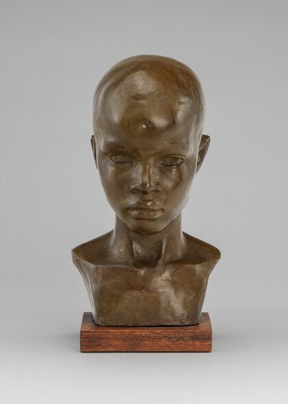 This free-standing, bronze-colored sculpture shows the head, neck, and the center of the collarbone of a young, bald boy. In this photograph, the boy faces us with his chin slightly lowered. He looks out from under a projecting brow. He has a flaring nose and his full lips are closed. The hollow of his throat is deep, and light glints off the tendons to each side. His chest is cropped to either side of his neck to create a trapezoidal shape just below his collarbone. The sculpture sits on a thin wooden base. The background is fog gray.