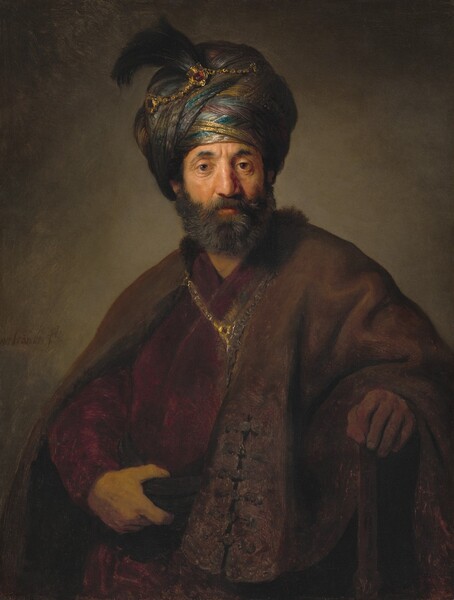 Shown from the waist up, a bearded man with a tanned, peachy complexion wears a turban and voluminous robes and looks out at us in this vertical portrait painting. Lit strongly from the left, the man’s brow is slightly furrowed over brown eyes, and his gray beard is trimmed. His turban is accented with a gold chain and feather, and his fur-lined robe is clasped over a burgundy-red garment with another gold chain. His right hand, on our left, grasps a sash that wraps around his waist, while his other hand rests on a wooden staff. The gray background behind the man deepens into shadows at the corners. The artist’s signature is painted to our left near the man’s upper arm. The first letter “R” is missing so it reads “embrandt ft.”