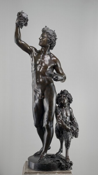This free-standing bronze sculpture shows a nude, cleanshaven man looking at a bunch of grapes he holds up in one hand as a faun, a boy with goat’s legs, looks on. In this photograph, the man’s lean, muscular body is angled to our left, and we see his face in profile. He has a straight nose, and his lips curl up in a smile. A leafy vine wraps around his thick, wavy hair, and another bunch of grapes hangs at the back of his neck. He holds the other bunch of grapes up high in his right hand, to our left. He holds a shallow dish in his other hand, with that bent elbow tucked near his ribcage. He stands with most of his weight on his left leg, to our right, so his hips and shoulders gently twist upward to the raised hand. The toes of his other foot rest behind him, his heel raised. To our right, the child-like faun comes up to the man’s hip. He has chubby cheeks and a small nose but muscles are defined in his torso and arms. He also has grape leaves on his head, with clusters of grapes at each ear. Wide-eyed and with an open smile, he gazes up toward the bowl and grapes that the man holds. A fabric strap is knotted on the faun’s right shoulder, and it runs diagonally across his body to his left hip where he holds two more large bunches of grapes and leaves. His upper thighs are covered with dense, wavy hair, while his lower legs are mostly bare, ending in cloven hooves. The pair stand on a circular base with bunches of grapes around the faun’s feet and hanging over the edge. The bronze is dark brown with bright white reflections where the light falls across the smooth surfaces. Touches of muted green patina are visible in some spots. The entire piece stands on a square, stone base marbled with tan and cream white, and the background is light gray.
