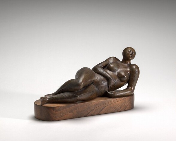 Cast from brown bronze, a stylized, nude woman reclines on an oval-shaped, wooden platform in this free-standing sculpture. In this photograph, the base is angled away from us so her head, to our right, is farther from us than her feet. She has a round, bare head with slits for eyes, a low ridge for a nose, and her lips are set in a straight line. She leans along the left side of her body. Her right arm drapes down across her midsection, and her thighs are stacked, with the foot of her top leg resting behind the bottom foot. Her features, including her arms, legs, and breasts, are rounded and smooth. The surface of the bronze is slightly textured throughout, and light glints off of the warm brown patina.
