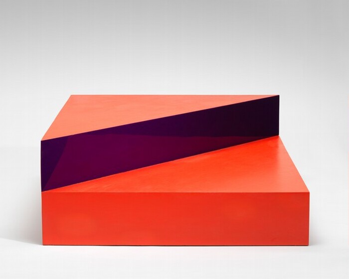 This abstract sculpture is made up of a square, scarlet-red box like a platform topped with a triangle, half the size of the square. In this photograph, we look onto one side of the square base, and the triangle lines up with the half farther from us. The front face of the triangle is a reflective, amethyst purple surface that reflects the square box beneath and the top surface appears to be coral orange. The backdrop behind the sculpture fades from light gray along the top of the photograph to white along the bottom.