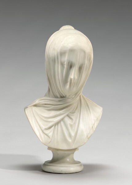 A free-standing, white marble sculpture shows the head and upper chest of a woman whose face is covered by a light veil. In this photograph, the chest is squared toward us, and the woman looks down and to our right. The veil has vertical folds over her face but we can still make out the hollows of her eyes, her straight nose, and her small mouth. Her wavy hair is pulled loosely back into a bun at the back of her crown. The veil gathers on either side of her face and crosses over neck to drape over her chest. The sculpture curves down in a wide U from near her shoulders, and is supported on a white marble pedestal. The background behind the sculpture is pale gray in this photograph.