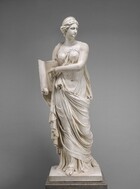 A woman carved from cream-white marble stands holding an open book to which she points in this free-standing sculpture. In this photograph, her body faces us but her head is turned to our right. She has a long nose, small mouth, and rounded chin. Her wavy upswept hair is topped by a semi-circular headdress. Her voluminous gown clings to her torso, leaving one breast exposed. The fabric falls in deep folds around her legs and sandalled feet. Her left shoulder, on our right, is bare. A band crosses her chest from that shoulder to her waist. She holds the tall book in her right hand, on our left, braced against that hip. Her other hand points to the words inscribed in Latin on its pages. It reads, “CALLIOPE REGI NA,HOMINVM, DIVVMQVE VO LVPTAS CARMINIS HE ROI NVMERIS FVLGENTIA SIGNIS AGMINA, BEL LANTVMQVE ANIMOS, ET P RAELIA CAN TO, INCLYTAQVE AETERNAE COMMI TO. NOMINA FAMAE.” She stands on a square base supported on a fawn-brown, veined marble plinth.