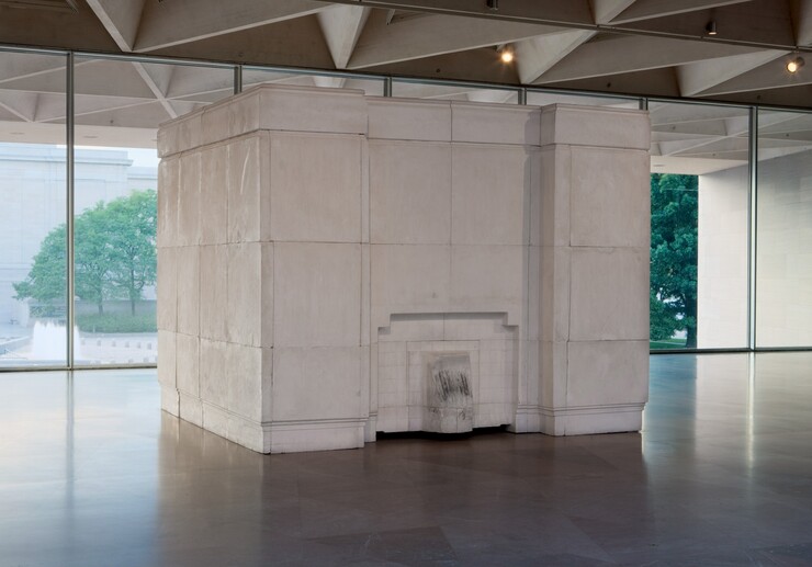 Set in a gallery against a row of windows, a free-standing, white plaster cube, nearly reaching the ceiling, angles back and away from us slightly to our right. The cube is made of blocks that fit together so the seams are visible. The side to our left is smooth. A short, tongue-like projection at the bottom center of the face to our right is surrounded by an inverted, squared off U arching over it. Though not obvious in this photograph, the artist created this work by filling an empty room with plaster. The tongue-like shape is the inside of a fireplace, and is lightly blackened where it lined the interior. The inset paneling in the cube would have been the room's mantlepiece, and inset bands along the floor and top of the cube would have been the baseboards and crown molding. The cube sits on a polished stone floor, and the ceiling of the room in which it sits is made of hollow triangular coffers. Opposite us, trees and a building are seen through the wall of windows behind the cube.