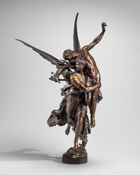 A winged woman has hoisted a young, muscular man up and across one shoulder in this freestanding bronze statue. In this view, the woman runs toward us, her back foot lifted, as she wraps her arms around the man’s thighs. She looks off to our left, her hair swept up. Long, narrow wings slice into the space behind her. Her chest and shoulders are covered with gold armor, and drapery swirls around her legs. The man is nearly nude, covered only with a cloth across his groin. His arms are flung wide, one reaching high in the air and the other holding a broken sword by the woman’s shoulder. His head lolls to one side, his lips parted. A laurel branch lies at the woman’s foot on a small mound textured like a rock. An inscription on the front rim of the base reads “GLORIA VICTIS.”