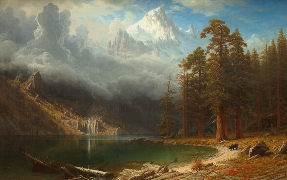 We look across the gleaming, forest-green surface of a lake at the foot of tall, jagged mountains in this horizontal landscape painting. The lake is flanked by a steep, gingerbread-brown hill on the left and a shoreline that forms a backward C curving from the lower center around and back along the right side of the composition. Strong sunlight from the upper left illuminates the center of the left-hand hill and the shoreline. The ground to our right is carpeted in rust-red, sage-green, and tan growth and is dotted with boulders. Tall trees with rust-brown trunks, crooked branches, and narrow canopies of caramel-brown and olive-green leaves fill the far end. Closest to us are dead tree trunks jutting out of the water and or lying on a flat, rocky outcropping nearby. Beyond the outcropping is a small black bear wandering down a sliver of sand-colored ground at the water’s edge. The hill on the left is covered with vertical rows of upright jagged boulders and slender, dark green trees marching up its slopes. A narrow, artic-blue waterfall cascades down its right side to empty into the lake. A thick layer of towering blue-gray clouds rises over the hill and lake, stretching back to the looming, snow-covered peaks that nearly brush the top edge of the canvas. The sky around the peak is vivid blue, scattered with high white clouds. The artist signed the lower right, “ABierstadt” with the A and B joined as a monogram.