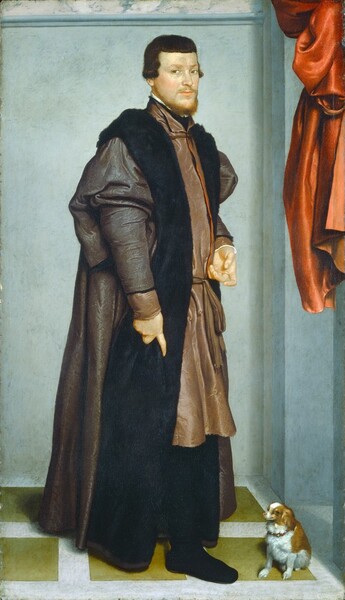 A light-skinned man with dark hair and a russet-orange beard stands wearing a long, voluminous, fur-lined coat next to a small dog near the corner of a room in this vertical portrait painting. His body is angled to our right but he turns his head to look at us with light brown eyes under faint, ginger brows. He has a straight nose, his cheeks are lightly flushed, and his light peach lips are closed. His ginger beard and mustache are closely trimmed. His wavy, brown hair reaches his ears, and short bangs skim across the top of his forehead. His knee-length, taupe-brown tunic is tied at the waist and has a high neck that encircles layers of black and white collars underneath. The floor-length, flint-gray coat, worn over the tunic, has puffed sleeves that gather at the elbows, and is lined with a broad band of black fur around the neck and down the front. With his right hand, to our left, he grips the fur lining with his index finger extended. His other hand is held in front of his waist with the palm facing up and fingers loosely curled. He wears black pants or stockings over black shoes. The small, long-haired dog sitting to our right near the man’s feet has caramel-brown spots along its back, around its eyes, and over its furry ears. It looks at us with dark eyes, and its mouth curves up. It comes about halfway up the man’s shin and wears a red collar with silver bells. The floor is patterned with fern-green squares in a white grid. The wall behind the man is pale blue. Marble molding along the top is white veined with gray, and a red curtain is bunched up in the upper right corner of the painting, presumably tied against the wall there.