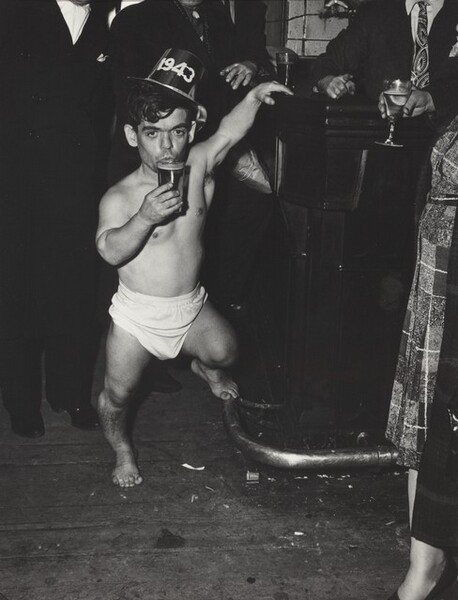This vertical, black and white photograph focuses on a dark-haired, barefoot man of short stature wearing a diaper and party hat, which bears the year, 1943. His left hand, to our right, reaches overhead and rests on a bar. His left foot perches on the footrail, and he holds a full glass to his pursed lips. He is surrounded by a black field, which eventually becomes evident as people wearing dark suits, their heads cropped by the top edge of the image.