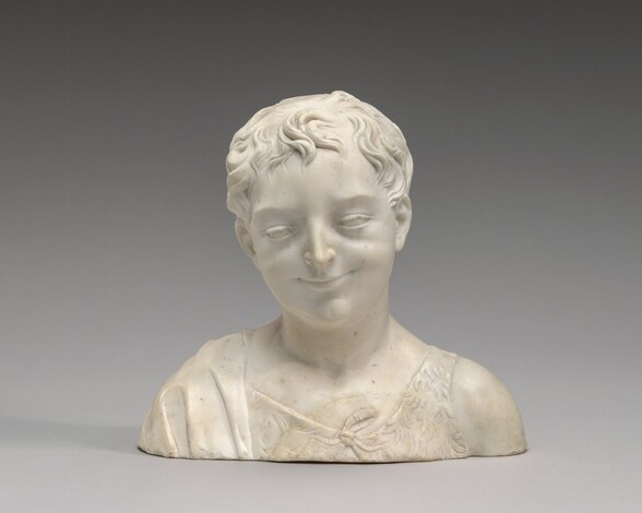 The head and shoulders of a young, smiling boy are carved from cream-white marble in this free-standing sculpture. In this photograph, his shoulders face us, and he looks slightly down and to our left. He has short, wavy hair that curls onto his forehead, and his skin is smooth. He has a short, straight nose, and his closed lips are turned up in a smile. He has rounded cheeks, and his chin has a shallow dimple. He wears a textured garment resembling fur and a swath of fabric falls over his right shoulder, on our left. The background behind him lightens from dark gray along the top to smoke gray along the bottom edge of the photograph.