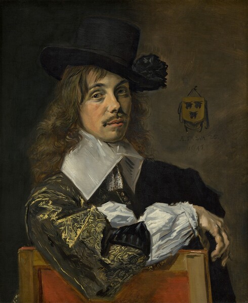 Shown from the waist up, a light-skinned man sits sideways in a wooden chair so the arm closer to us rests along the chair’s back in this vertical portrait painting. Brushstrokes are visible throughout, especially in the clothing and hair. The man’s right arm, to our left, drapes across the back of the chair. His face turns slightly to our right, and he looks at us with gray eyes under black, curving brows. He has an oval face with high cheekbones and a snub nose. His coral-pink lips are closed and framed by a mustache and pointed patch of hair under his lower lip. He is lit from our left so the far side of his face is in shadow. His loose, curly brown hair cascades over his shoulders. He wears a tall black hat with a stiff, wide brim with a black rosette or pompom on the brim to our right side. The hat is tipped slightly to one side so it angles away from us. A flat, stiff, white collar extends from his chin down to his shoulders, and two tassels hang at this throat. The voluminous sleeve along the back of the chair has a gold pattern picked out in loosely painted, swirling, feathered, and flaring lines against a black background. The jacket is unbuttoned along the forearm to reveal a stark white undershirt that is gathered with a narrow ruffle at the wrist. The chair has a crimson-red center framed by wooden rails to each side and along the top. The background is earth brown to our right of the man, and deepens to dark shadows along the left. A coat of arms, the size of the man’s palm, hangs from the back wall. Three black cow heads create an inverted triangle on a gold field. Under the coat of arms the artist inscribed, “AETA SVAE.22 1645.”