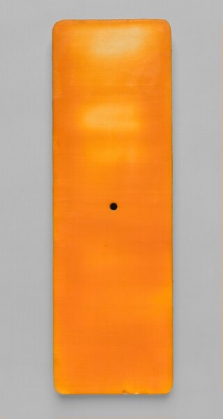 This rectangular, tangerine-orange panel has rounded corners and a black dot at the center. It hangs on a dove gray wall and is about three times as tall as it is wide. The surface appears streaked with golden yellow where light reflects off some areas of the wax, especially in the top half.