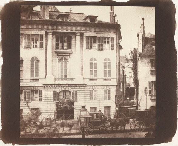 We look across a street at the façade of a five-story building with a pitched roof in this square sepia-toned photograph. Vertical rectangular windows are spaced along the lowest two levels of the building, which appears to be constructed with large rectangular stones. There is a squat, arched window over a dark opening at the bottom center. The façade of the next two stories is smooth. Tall, narrow arched windows line the third story, and the fourth story has smaller rectangular windows with shutters. Columns frame the central bank of windows in the third and fourth stories. Dormers of the fifth level protrude from the low pitched roof, which nearly reaches the top edge of the photograph. Another house is visible through a narrow alleyway to our right, and another shorter house is cropped by the right edge. Some details of the three horse-drawn carriages lining the street in front of the houses are blurry. The image is surrounded by a wide black band with uneven edges.