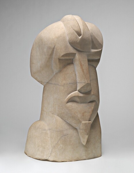 This free-standing marble sculpture shows a man’s abstracted head and shoulders carved in angular planes. The top third of the piece is a smooth head, perhaps bald, with a curling, pointed oval shape coming down the middle of his forehead. The middle third is the elongated, rectangular face. Closed eyes are suggested by thin ridges carved in deep recesses on either side of a long nose, which is shaped by flat planes. The mouth is delineated by a thick, downturned, upper lip, and a narrow goatee hangs from the pointed chin. The bottom third is the shoulders, suggested by a downward-facing arch carved on the side we can see. The surface is rough and unpolished with a few streaks of gray in the sand-colored marble. This photograph shows the sculpture angled to our right against a pale gray background.