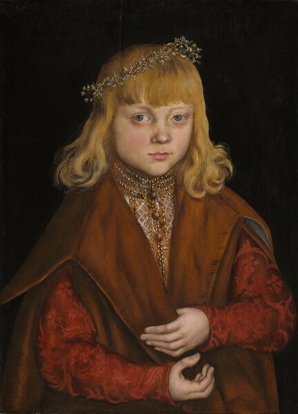 Shown from the waist up, a young boy with blond hair faces us, wearing an embroidered garment and heavy cloak in this vertical portrait painting. His body angles slightly to our right, but he looks just off to our left with pale blue, faintly bulbous eyes. He has pale skin, flushed cheeks, a delicate nose, a pointed chin, and his pink lips are closed. His wavy hair curls around his shoulders, and a fringe of bangs spans his forehead. A crown of black pearls, gold, and green stones is set at an angle on his head. His high-necked light brown shirt is edged with gold and embroidered with white and gold. The long sleeves are brocaded with a ruby-red floral pattern against darker garnet red. He holds the edge of a caramel-brown, sleeveless overcoat with one pale hand, and the other is cupped with fingers pointing upward just below. He is shown against a black background.