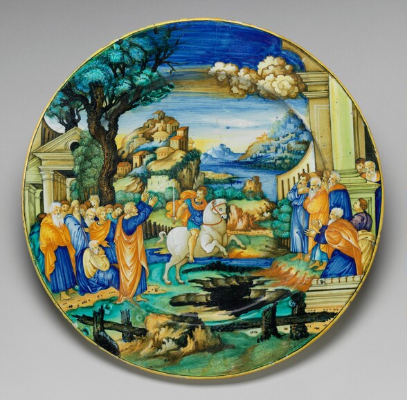 This circular earthenware plate is painted with a landscape scene showing a man on horseback in the center flanked by a group of about a dozen men to the left and ten boys and men to the right. The white horse faces our right as it rears up on its back legs and looks down at a dark chasm in the earth beneath its front legs. The man astride the horse has pale skin and light brown hair, and he holds a sword as he looks back to the crowd to our left. Everyone in the crowd also has pale skin. Most of the people wear vibrant, royal and sky blue and pumpkin orange, with a few touches of butter yellow, turquoise, and purple. Both groups are situated in front of sandstone-colored columns and pediments, which flank the scene. A cluster of buildings rises along a ridge in the middle distance to the left of center, and more structures are built along a riverbank to our right. The river weaves away from us to blue mountains in the deep distance.