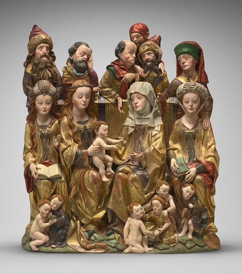 Four women, six men, and seven babies and children are gathered in three rows in this freestanding, painted wooden sculpture. The people all have pale, peachy skin with rosy cheeks, round faces with double chins, and thin, curving brows under high foreheads. They mostly wear gold robes over garments in gray, dark blue, plum purple, spruce green, or red. The four women sit hip-to-hip across the middle of the grouping. The two center women angle slightly in toward each other. To our left in that pair, Mary holds a nude baby Jesus on her lap. Mary’s long, loose, light brown hair is held back with a twisted band. The chubby Jesus on her lap reaches for the woman to our right, Saint Anne, who wears a voluminous wimple that covers her face, chin, neck, and shoulders. The women to either side of the central pair hold books, one open and one closed, and wear turban-like headdresses over long, wavy hair. Six children play together in two groups at the women’s feet. Some are nude and others wear dark or gold robes. A pair eat grapes to our left. The right-most child rides a hobby horse and holds onto the hand of the woman behind him with his tiny hand. In the top row, a man stands behind each of the four women except for Saint Anne, who has three men standing over her shoulder. Two of the six men are cleanshaven, and the others have beards. Some are bareheaded and others wear turbans or a cap. Their faces are lined, and they look down or off into the distance, most under furrowed brows. The gold on the robes is tooled to create patterns.