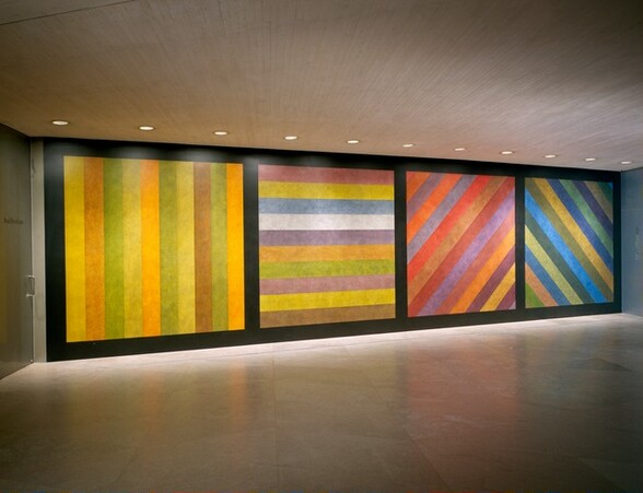 A long wall is painted with from floor to ceiling with black borders to create four squares, each of which is filled with colorful, vertical, horizontal, or diagonal stripes. The stripes are all the same width, and are painted in slighly mottled colors to create a sense of texture. In this photograph, we are to the left so the wall moves a bit away from us to our right. The ceiling and floor are both light pink stone or concrete, and a row of recessed lights in the ceiling near the wall shines lights onto the paintings there. The left-most square has vertical stripes in daffodil yellow, amber, gold, laurel green, and one caramel-brown stripe. The second square has horizontal bands. Along the top is a stripe of dusty rose, and then stripes in lemon yellow, faded blue, white, muted, amber, laurel green, mauve pink, another lemon yellow, and caramel brown. The third square has diagonal lines that reach from the bottom left corner to the upper right. These stripes are in warm shades of pumpkin orange, lavender, light brown, scarlet, brick red, mauve, dusty rose, and plum purple. The fourth square has diagonal bands going in the opposite direction, from bottom right to top left. Those stripes are in cool tones of denim, steel, and azure blue; pine, sage, and spring green; and peanut brown.