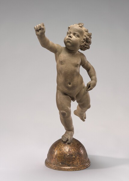 Sculpted in clay, a toddler balances his plump body on his right foot as he raises his left leg and right arm. In ths photograph, his body is squared to us. His left hand, on our right, rests near his hip. Gazing up and to our left, his cheeks are puffed in blowing through pursed lips, and his hair falls in soft ringlets. The fingers of his raised hand are folded into a loose fist. Tipped slightly forward onto his toes, he balances on a gold-half dome.