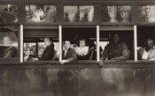 This horizontal black and white photograph captures the exterior of a trolley car cropped to show only the mid-section with six passengers inside. The passengers look out of five windows on the left side of the trolley, so their bodies face our left in profile. The four people at the front end, to our left, are white; two people at the back, to our right, are Black. From left to right: a man looks out through the glass of the closed window along the left edge of the photograph. The remaining windows are open. An older woman wearing a dark coat looks at us from under arched eyebrows, lips pursed, out of the next window, to our right. Two children—a boy of about eight and a girl of about two—look out of the central window. Closer inspection reveals the dark form of their caregiver lost in the shadowy interior of the trolley. A Black man wearing a long-sleeved, button-down shirt leans onto the window ledge with both forearms in the next window, and a woman wearing glasses looks over her shoulder, up and away from us in the right-most window, which is cropped by the edge of the photograph. The scene behind the photographer is reflected in glass panes above the seats on the exterior of the trolley.