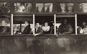 This horizontal black and white photograph captures the exterior of a trolley car cropped to show only the mid-section with six passengers inside. The passengers look out of five windows on the left side of the trolley, so their bodies face our left in profile. The four people at the front end, to our left, are white; two people at the back, to our right, are Black. From left to right: a man looks out through the glass of the closed window along the left edge of the photograph. The remaining windows are open. An older woman wearing a dark coat looks at us from under arched eyebrows, lips pursed, out of the next window, to our right. Two children—a boy of about eight and a girl of about two—look out of the central window. Closer inspection reveals the dark form of their caregiver lost in the shadowy interior of the trolley. A Black man wearing a long-sleeved, button-down shirt leans onto the window ledge with both forearms in the next window, and a woman wearing glasses looks over her shoulder, up and away from us in the right-most window, which is cropped by the edge of the photograph. The scene behind the photographer is reflected in glass panes above the seats on the exterior of the trolley.