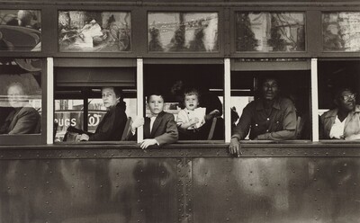 This horizontal black and white photograph shows the exterior of a trolley car cropped to show only the mid-section with six passengers inside. The passengers look out of five windows on the left side of the trolley, so their bodies face our left in profile. The four people at the front end, to our left, are white; two people at the back, to our right, are Black. From left to right: a man looks out through the glass of the closed window along the left edge of the photograph. The remaining windows are open. An older woman wearing a dark coat looks at us from under arched eyebrows, lips pursed, out of the next window, to our right. A boy and a young girl look out of the central window. Closer inspection reveals the dark form of a woman next to them, lost in the shadowy interior of the trolley. In the next window, a Black man wearing a long-sleeved, button-down shirt leans onto the window ledge with both forearms, and, in the right-most window, a woman wearing glasses looks over her shoulder, up and away from us. That last window is cropped by the edge of the photograph. The scene behind us is reflected in glass panes above the seats on the exterior of the trolley.
