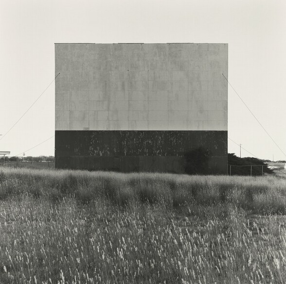 We look across a stretch of softly waving long grass at a drive-in movie screen tethered to the ground with long guy wires in this square black and white photograph. The base is dark under the lighter panels that make the screen. The horizon comes just under halfway up this composition, and it lines up with the foot of the screen so the whole is outlined against a blank sky.