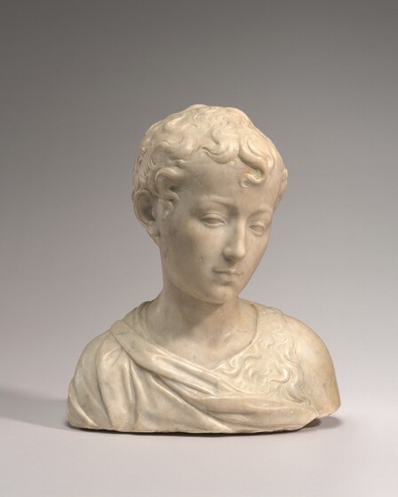 The head and shoulders of a young boy with short, curly hair is carved from cream-white marble in this free-standing sculpture. In this photograph, his shoulders are squared to us, and he looks down and to our right. He has delicate features and smooth skin. Fabric wraps around one shoulder and across his chest, and a wide strip like animal fur goes over the other shoulder, to our right. The sculpture is cut straight across the chest just below the shoulders, and it sits in front of a background that lightens from smoke gray along the top to nearly white along the bottom.