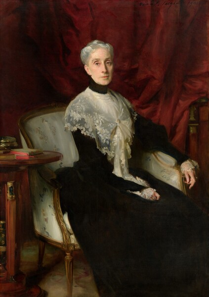 A woman with pale skin, wearing a sheer, lace shawl over a high-necked, ink-black dress sits in an upholstered chair in front of a dark, cranberry-red curtain in this vertical portrait painting. The chair and her body nearly fill the composition, though her feet may be cropped by the bottom edge. She sits angled to our right but she turns her face to look at us with gray eyes under arched brows. Shadows line the creases above her eyelids. Set in her thin, oval face, she has a long nose, high cheekbones, and her coral-red lips are closed in a straight line. She is lit from our left, and the light glints in her silvery-gray hair, which is pulled up and back. Her velvety-black, long dress seems to have puffed upper arms over long sleeves that end with wide lace cuffs. The lace shawl wrapped around her shoulders is fastened with a gold pin at her chest, and the ends fall into her lap near her right hand, closer to us. Her other arm rests along the arm of the low-backed chair so her long, elegant fingers dangle off the end. She wears a ring on each hand, including one glinting turquoise, perhaps indicating that it is a dark stone, and one with a pearl. The fabric on the chair is loosely painted with a tan-colored floral design against oyster white. A small, wooden table with a round top to our left, near her right elbow, holds a book with a sage-green cover and vivid red edges, and a shiny, gold, round box. Barely visible against the red curtain behind the woman in the upper right corner is the artist’s signature and date: “John S. Sargent 1901.”