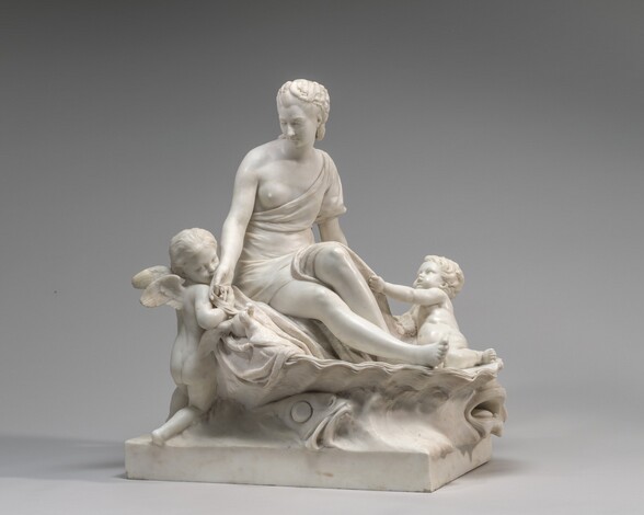 A seated woman with upswept hair is flanked by two chubby male children in this free-standing, white marble sculpture. In this photograph of the sculpture, the woman’s body faces us, and the rectangular base on which they sit is angled slightly away from us to our left. The woman sits in half of an oversized, upturned clam shell with her legs angled to our right. Her near leg is outstretched while the other bends at the knee. Her toga-like garment leaves her near breast exposed and wraps around her back to cover her lower torso and lap. She looks down to the winged boy closest to us, to our left, who half-stands and half-kneels at her side. He holds her hand and kisses the back of it, as he turns his face toward us. Two doves near the boy nestle in the drapery on which the woman sits, their beaks interlocked. On the woman’s other side, to our right, the second boy sits in the shell by her feet. He leans back to look up at her with his mouth open, as he grabs her garment with one hand. Two sea creatures with gaping mouths support the undulating curves of the shell.