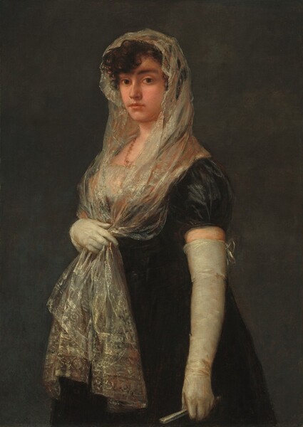Shown from the knees up against a dark gray background, a young woman with peachy skin stands with her body angled to our left and she looks at us with brown eyes in this vertical portrait painting. She has dark, arched brows, a straight nose, and her full, coral-pink lips are closed. Her dark brown, curly hair is covered by a mantilla, an ivory-white, lacy scarf highlighted with pale gold and subtle, russet-red highlights. She holds the two long ends of the mantilla with her right hand, farther from us, below her bust. The ends of the scarf fall to mid-thigh over her long, ink-black dress. The bust of her dress is also white and shimmers, like the mantilla. Long, tight-fitting, off-white gloves nearly reach the short, puffed, cap sleeves of the dress. The back of the glove on her left arm, closer to her, is tied at the top with a bow, and she holds a silvery-white, closed fan in that hand. A brownish-coral colored necklace is painted as a squiggle of paint around her neck. Barely visible, the painter signed the painting in the lower left: “Goya.”