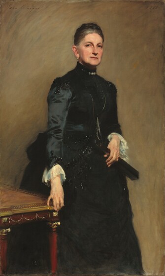 Seen from the shins up, a woman with pale skin, iron-gray hair bound in a low, wide knot atop her head, and wearing a high-collared, long-sleeved black dress, stands next to a table against a caramel-brown wall in this vertical portrait painting. Her body is angled to our right and she looks out at us from the corners of her eyes with dark eyes under arched brows. She has a straight nose and slight jowls along her jaw line. Her thin, coral-red lips are set in a straight line. A round pearl hangs from the earlobe we can see and glints in her hair suggest pearls or other ornaments. Three jewels, also perhaps pearls, glisten at her high collar and black beads catch the light at the shoulders and cuffs of the puffy, long sleeves. The bottom edge of the tight-fitting bodice also seems to be beaded, and the full skirt flares out at the hips. The sleeves end with white, ruffled cuffs. She holds a closed black fan with her left hand, on our right, and wears a gold ring on that pinky finger. She stands just behind the corner of a cherry-red table edged with gold, to our left, and grips the corner with the fingertips of her opposite hand. The portrait is painted with loose but blended brushstrokes, giving it a soft look. The artist signed the painting in the upper left corner, “John S. Sargent” with the date in the upper right, “1888.”