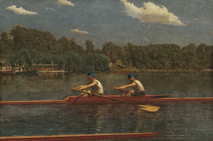 Two men with pale skin, wearing white sleeveless shirts and royal-blue caps, row in unison in a long, narrow boat on a placid blue river in this horizontal landscape painting. The low, honey-colored wooden boat extends off both sides of the canvas. Both men face our left as they row to our right. The man in front looks ahead of him, beyond the stern of the boat, and the man to our right, closer to the bow, tucks his chin down to look past his shoulder. Their bare, muscled arms are extended straight as the two oars sweep back. The tip of another boat runs close and parallel to the bottom edge of the composition, spanning the left three-quarters of the painting. The opposite riverbank is lined with a dense forest of pine-green trees. People crowd along the decks of a steamboat and a paddleboat near the riverbank to our left. Another narrow skuller, rowed by four people wearing ruby-red shirts, cuts through the water at the back center of the river. The riverbank beyond is lined with people, painted with strokes of black and white, and miniscule touches of red. The horizon comes halfway up the composition. Cream-white clouds float across a muted, topaz-blue sky above the trees.