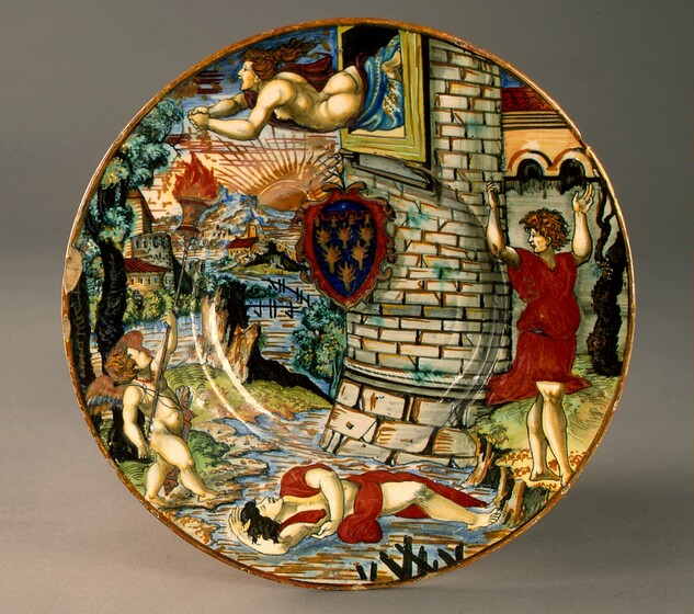 This circular plate is painted with an almost cartoon-like scene showing four people with pale skin around a stream, near the exterior of a tower. At the top of the plate, a nude woman is shown from the thighs up as she falls horizontally from an open window at the top of the tower. Her back, buttocks, and a single breast are exposed to us while a deep red and blue fabric wraps over her far shoulder and around her thighs. Her hands clasp as she reaches her arms forward, and her copper-red hair flies behind her. Her mouth is wide open. Directly below her, at the bottom center of the plate, a man partially wrapped in ruby-red drapery lies on his back in the stream. One arm is flung over his head and the other hangs across his body. He has black hair, and his eyes are closed. On the right side of the plate, a second man stands on the bank of the stream with his body facing us, as he turns his face to our left in profile. He has curly, auburn brown hair, and the eyebrow we can see is furrowed. He wears a knee-length red tunic, and he raises both arms high overhead. Finally, the fourth winged, child-like person stands on the opposite bank, to our left. He is nude except for a quiver of arrows slung across his chest. His babyish body is chubby with small wings between his shoulder blades. He faces the center of the scene and looks up at the woman while raising a long torch toward her. The tower is made of parchment-white stones outlined with mustard yellow and black. A shield or coat of arms hangs or hovers just above the center of the plate. It has a rounded top and pointed bottom, and has six gold, tree-like forms against a navy-blue background, all within a scrolling, red frame. The stream winds into the distance between buildings along the banks. Cobalt-blue and sepia-brown mountains line the horizon, and a sepia-brown sun with linear rays sets behind them. The plate is edged with a thin band of golden brown, and there are several chips around the rim. The plate is shown in front of a light grey background and it casts a light shadow to our right.
