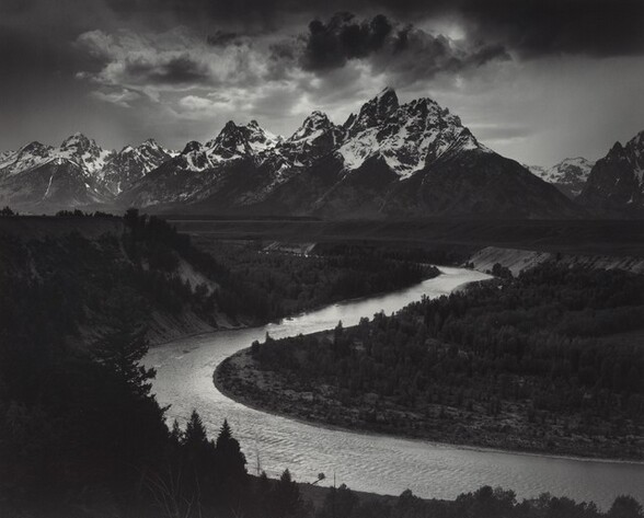 We look down into a darkened valley with a shimmering, curving river winding back to a screen of craggy, snow-topped mountains in the distance in this horizontal black and white photograph.