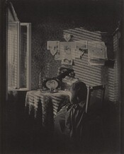 A woman with light skin wearing a long, corseted dress with a full skirt and a wide-brimmed hat with feathers sits writing or drawing at a round table next to an open window in this vertical, black and white photograph. Light coming in through the window to our left creates slanting stripes of light and shadow across the table and the wall opposite us, suggesting it filters through blinds or slats. The woman sits in a straight-backed, wooden chair with a caned back, angled away to our right almost in profile. She looks down and bends slightly over the paper and pen. Her hair is pulled back in a bun at the back of her head under the hat, and her dark dress has a wide lace collar. The table is covered with a patterned cloth and holds a tall candlestick with a twisted candle, an ink bottle, a small, lidded jar, and a picture of a woman in a frame decorated with daisies. On the wall opposite us six photographs or drawings hang on the wall. Two show landscapes, three show a woman or women, and one shows a man. Three heart-shaped decorations tied with bows hang among the other papers and a bird cage hangs to our right. The wallpaper has a dense, formal, stylized floral pattern. The windowpanes swing into the room to our left, and a sliver of an open doorway lines the right edge. The top and bottom zones of the photograph are swallowed in deep shadow.