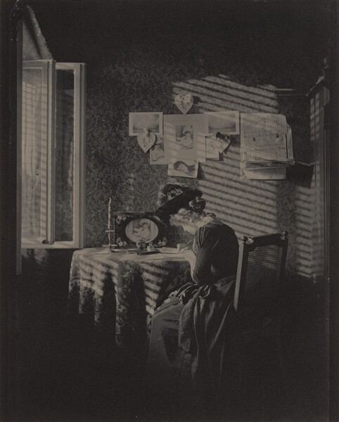 A woman with light skin wearing a long, corseted dress with a full skirt and a wide-brimmed, feathered hat sits writing or drawing at a round table next to an open window in this vertical, black and white photograph. Light coming in through the window to our left creates slanting stripes of light and shadow across the table and the wall opposite us, suggesting it filters through blinds or slats. The woman sits in a straight-backed, wooden chair with a caned back. We see the curve of her cheek and the tip of her nose as she bends slightly over the paper and pen. Her hair is pulled back in a bun at the back of her head under the hat, and her dark dress has a wide lace collar. The table is covered with a patterned cloth and holds a tall candlestick with a twisted candle, an ink bottle, a small, lidded jar, and a picture of a woman in a frame decorated with daisies. On the wall opposite us, six photographs or drawings hang on the wall. Two of them show landscapes, three show a woman or women, and one shows a man. Three heart-shaped decorations tied with bows hang among the other papers and a bird cage hangs to our right. The wallpaper has a dense, brocade-like floral pattern. The windowpanes swing into the room to our left, and a sliver of an open doorway lines the right edge. The top and bottom zones of the photograph are swallowed in deep shadow.