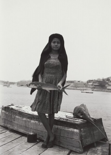 A dark-skinned girl stands on a dock holding a fish wider than her hips down in front of her with both hands, one ankle crossed over the other in this black and white photograph. Her body faces us, but she looks up and off to our right. A dark cloth covers her hair and shoulders over a knee-length dress. She wears loafer-style shoes. She stands in front of an upturned rowboat with a severed swordfish head laid on the flat stern, to our right. The long sword-like upper lip angles into the lower right corner of the photograph, and the fish’s large eye is a gaping hole. Buildings on a spit of land that comes about halfway up the composition enclose the body of water in the distance. The contrast of the girl’s dark head covering against the white sky creates an optical effect, making the cloth seem to glow.
