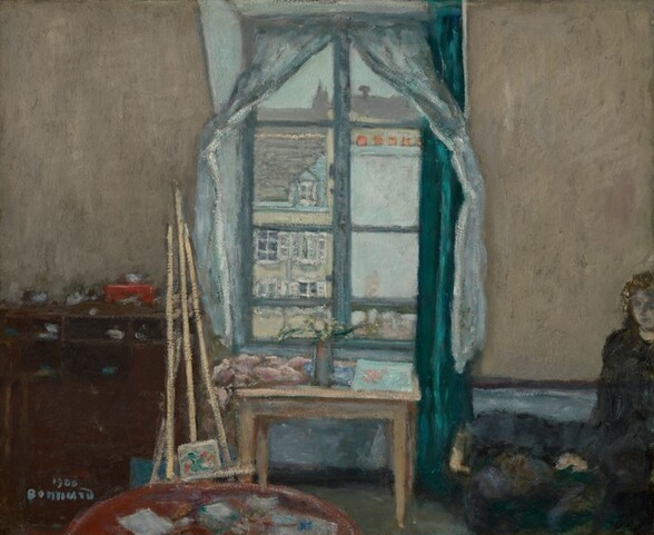 We face a wall lined with furniture to either side of a tall window that looks out onto buildings in this almost square painting. The scene is painted with blended and broad strokes, which makes our view a little blurry so many details are indistinct. The walls are tan with a thin, dark blue line above a wide band of lighter blue along the bottom of the wall. The window is in the center of the wall opposite us. A teal-blue curtain hangs along its right, and sheer, artic-blue drapes are parted and fastened to the wall on each side. The view out the window shows a few brightly lit, pale blue and yellow buildings with gray roofs. Under the window, a light wood table is scattered with objects, difficult to identify, and one pale turquoise square that could be a painting. More objects are cluttered on the dark wood bureau to our left and a round table closer to us, which is cut off by the bottom edge of the canvas. A delicate straw-colored easel holding a small canvas stands just beyond the round table. A girl sits in the lower right corner of the composition, and she is cropped by the right and bottom edges. Her skin is pale and tinged with smoky blue. She wears a wreath in her hair, presumably of flowers, and her long dress and the bed or chair on which she sits blend together in tones of navy, spruce blue, and forest green. The artist signed and dated the lower left, “1900 Bonnard.”