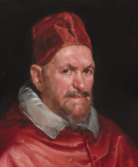 Shown from the shoulders up, a man with a flushed complexion, wearing a crimson-red cap and garment, looks at us from the corners of his eyes in this vertical portrait painting. He is brightly lit from behind us and the mahogany-brown background is deep in shadow. The man’s shoulders angle to our right and he turns his head slightly to look at us with brown, deep-set eyes under arched brows. His wispy, auburn-brown goatee is tinged in silver gray. His nose is slightly bulbous, and his thin lips are tightly closed in a straight line. Wrinkles and jowls give his face a paunchy look. His tall, flat-topped, fez-shaped hat covers his hair. A white, nearly translucent collar curls up along his neck and flares out to points at his collar. Rose-pink highlights on his red cape suggest it is a shiny material, like silk, and it is fastened with several small, dark buttons down the front.