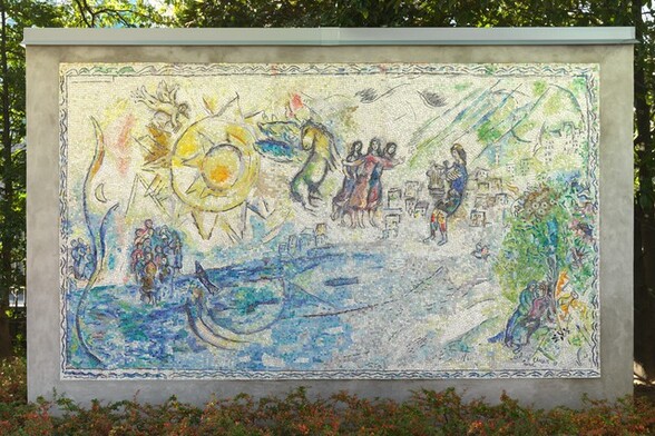 Created with tiny, colored stones and glass, this mosaic shows groups of people along and near the edge of a brilliant, topaz-blue body of water with a large, lemon-yellow, stylized sun in the sky in this horizontal composition. The aquamarine blue and vibrant yellow along with lime green, lilac purple, oyster white, and teal used in the people and landscape give this work a shimmering look. A pair of people recline together under a tree in the lower right corner. A sheep stands nearby, and a bird perches in the tree above. Beyond the tree and to our left, a cluster of box-like forms suggests a city in the distance. Larger in scale, so seeming closer to us, a man wearing a lapis-blue toga and a yellow cap holds a stringed instrument, a lyre, in front of his body. To the left, at the center of the composition, a trio of women each wearing amethyst-purple, magenta-pink, or lapis-blue dresses stand close to each other. They have long, dark hair and their bodies and garments are outlined in black. Between them and the large sun to our left, a winged horse with a buttercup-yellow body and turquoise-blue wings rears up. Near the upper left corner, the stylized sun is represented by a disk surrounded by pointed peaks, surrounded by a larger disk outlined with another ring of triangular points. A winged person holding a set of pan pipes flies above the sun to our left, over a small crescent moon below. Near the lower left corner, two groups of people gather around a body of bright blue water occupied by two fish. The artist signed and dated the work in the lower right corner: “MArC ChAgAll 69.” The mosaic is displayed outside in front of a screen of trees, with a band of bushes with green and orange leaves below.