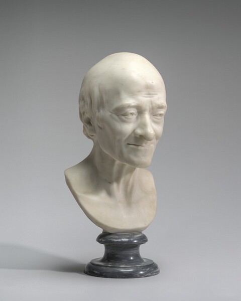 A white marble sculpture shows the head and upper chest of an elderly, gaunt, bald, smiling man. In this photograph, the man’s face is angled to our right, and he looks off in that direction with hooded eyes under bushy brows. His forehead, eyes, and mouth are lined with wrinkles. He has a bulbous, slightly hooked nose and high cheekbones. He smiles with his thin lips together. There is a fringe of hair over the ear we can see, and his head is otherwise smooth. The tendons on his neck stand out. The narrow base of the sculpture curves in a U down past his collarbones, and is supported on a charcoal-gray, polished marble base. The sculpture casts a light shadow against a pale gray background to our left in this photograph.